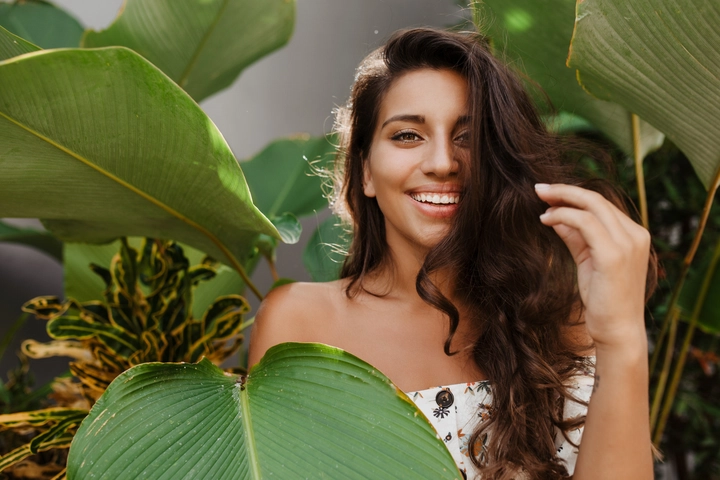 Long, healthy brown waves falling over the shoulders of a woman standing among lush tropical leaves, reflecting 2026 hair care trends focused on natural hair care, plant‑based ingredients, scalp health, and glossy, hydrated texture.