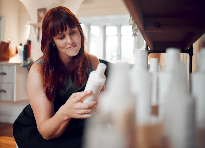 A woman examining a white bottle of private label hair care product among several options in a bright indoor setting.